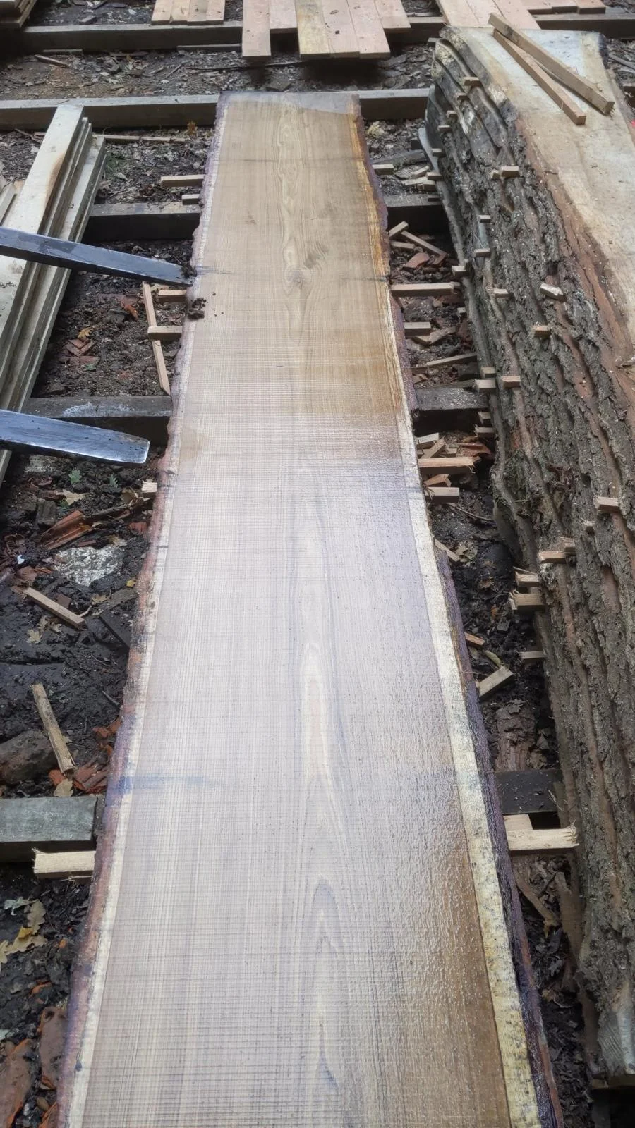 Long plateau brut posé sur le banc de sciage, à côté d’une pile de plateaux avec écorce
